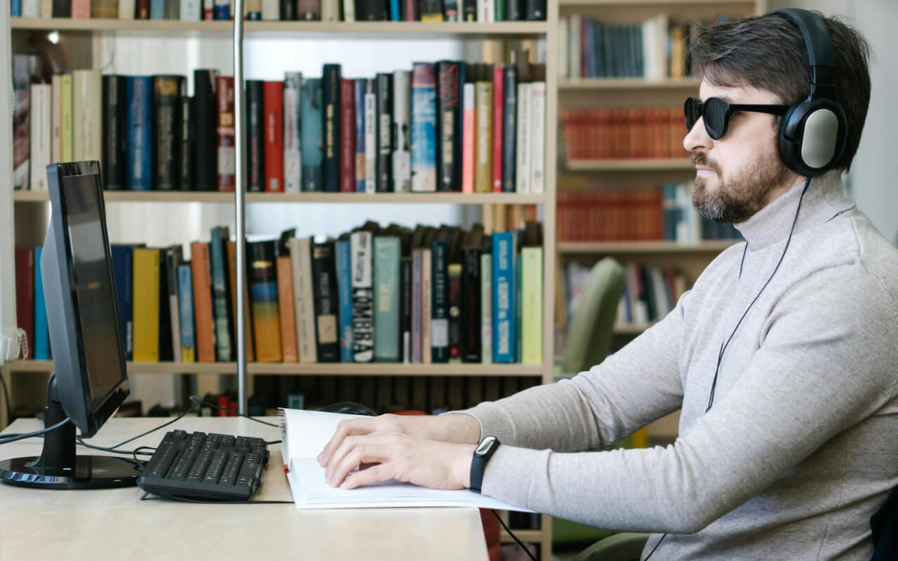 A visually-impaired man sits at a computer using a braille interface and wearing headphones.