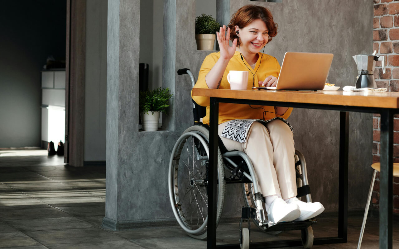 A woman in a wheelchair sits at a table using video chat on a laptop.